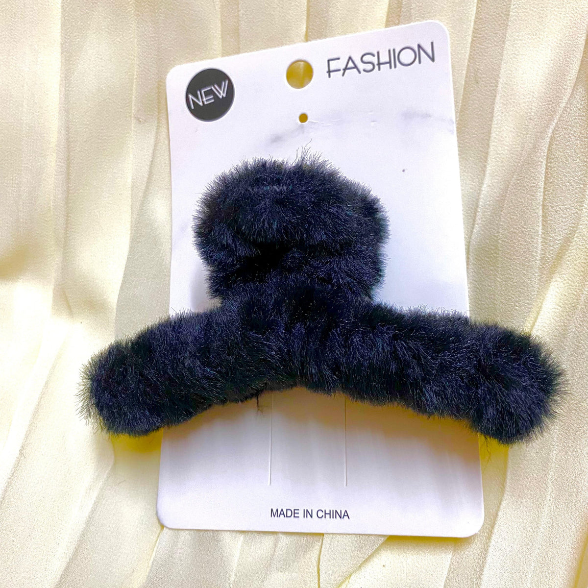 Navy blue fuzzy hair clip on a white card with 'New Fashion' text, against a beige fabric background.